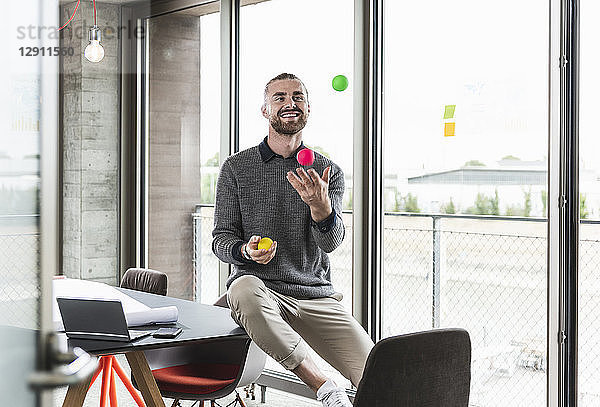 Smiling young businessman sitting at the window juggling with balls