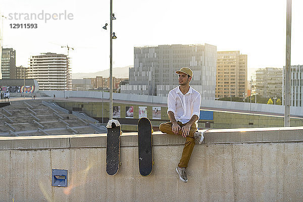 Young man sitting on urban wall next to skateboards at sunset