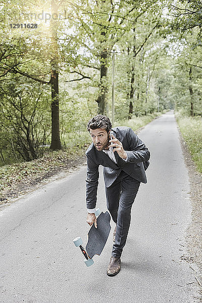 Businessman walking with skateboard and smartphone on rural road