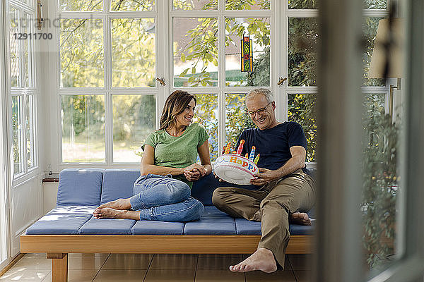 Happy mature couple sitting on couch at home with fake birthday cake