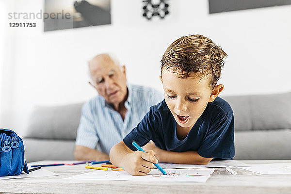 Portrait of little boy drawing with coloured pencils while his grandfather in the background watching him