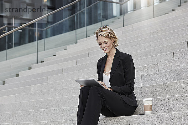 Blond businesswoman with coffee to goe sitting on stairs using tablet