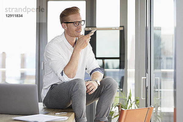 Smiling businessman sitting on desk in office using smartphone