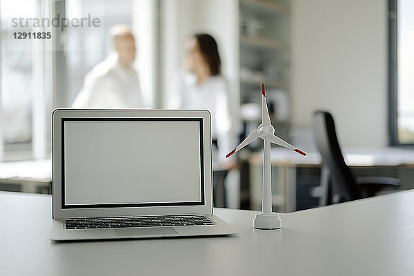 Laptop and model of wind wheel on shelf in office  with two people talking in background