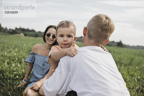 Brother consoling crying baby on a field with mother in background
