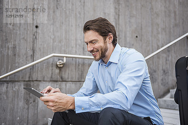 Smiling businessman with suitcase sitting on stairs using tablet