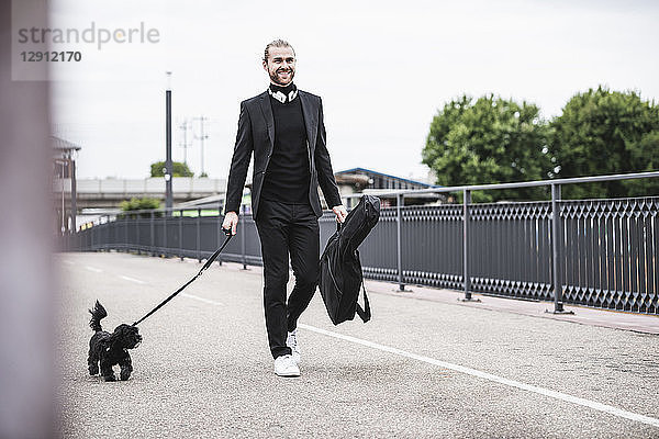 Fashionable young man holding guitar case walking with dog on a bridge