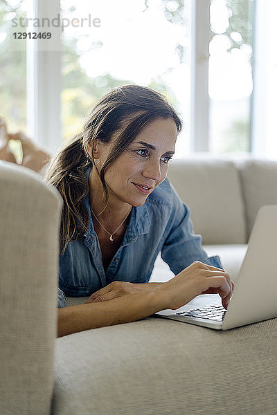 Smiling mature woman lying on couch at home using laptop