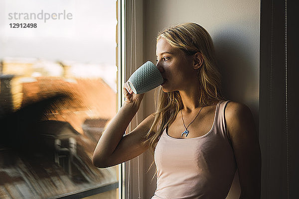 Blond young woman drinking coffee from mug looking out of window
