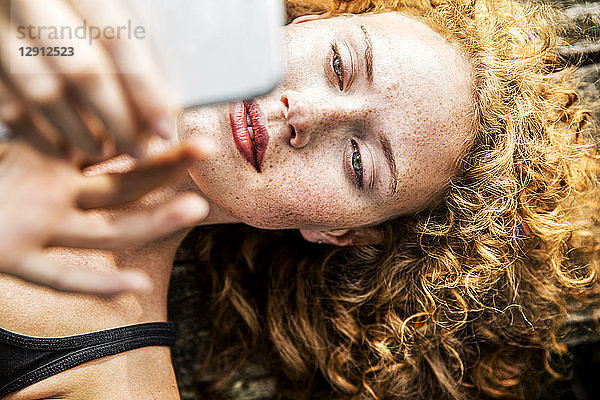 Portrait of redheaded young woman lying on bench using cell phone