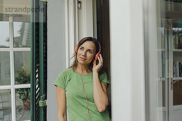 Mature woman standing at terrace door listening to music with headphones