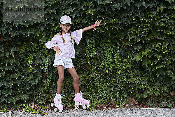 Cheerful little girl posing against ivy plants with pink roller skates in a park