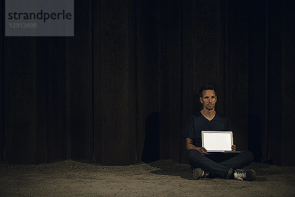 Mid adult man sitting cross-legged on ground  showing blank laptop screen