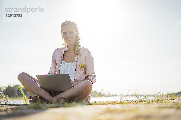 Portrait of smiling woman sitting at the riverside in summer using laptop