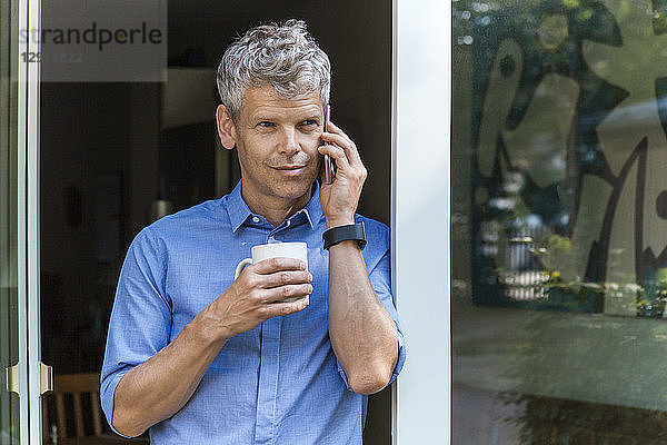 Portrait of mature man on the phone leaning agaich door case with coffee mug