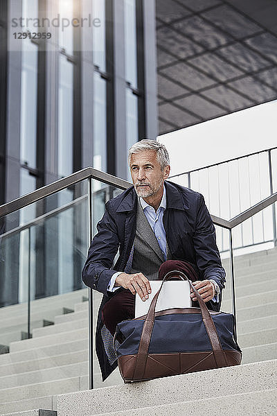 Portrait of mature businessman with traveeling bag and tablet crouching on stairs outdoors