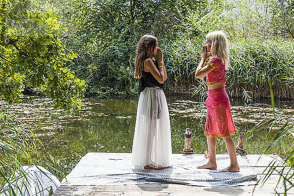 Two girls standing face to face on jetty at a pond in fancy dresses