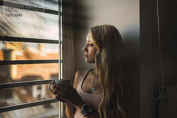 Blond young woman holding coffee mug looking out of window