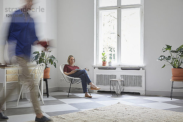 Mature woman reading magazine at home with man passing by