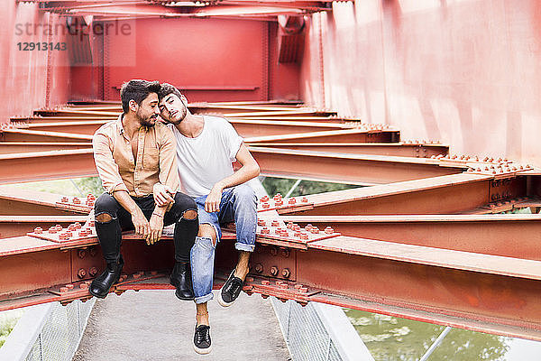 Young gay couple in love sitting on steel girder of a footbridge