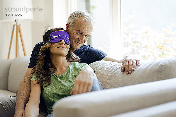 Smiling mature couple sitting on couch at home with woman wearing eye mask