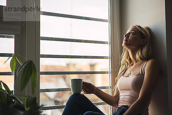 Blond young woman holding coffee mug sitting in windowsill