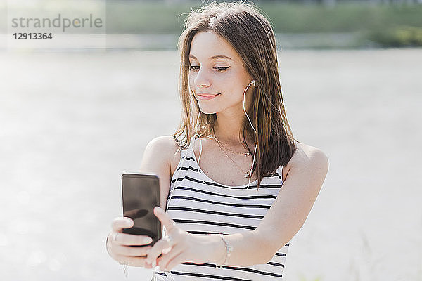 Smiling young woman with smartphone and earphones at the waterfront