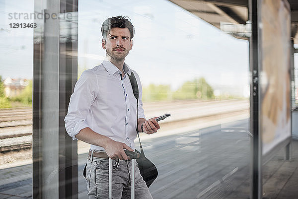 Businessman with baggage and cell phone standing on station platform looking around