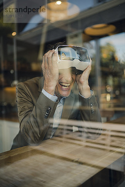 Mature businessman sitting in coffee shop  looking through VR glasses  laughing