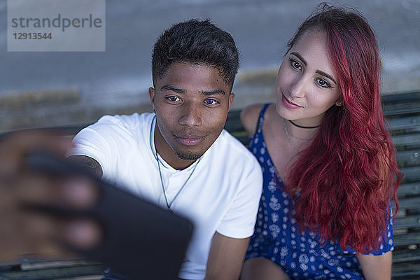 Portrait of young couple sitting together on bench taking selfie with smartphone