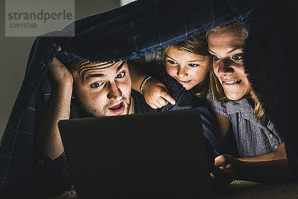 Happy family hiding under blanket  watching film on laptop