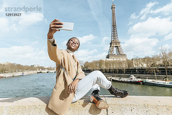 France  Paris  Woman sitting on bridge over the river Seine with the Eiffel tower in the background taking a selfie