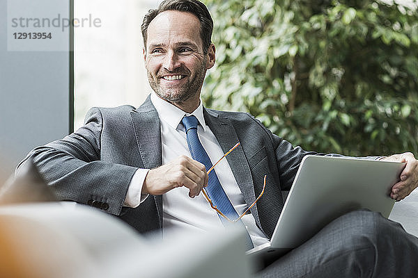 Portrait of smiling businessman sitting in lobby with laptop