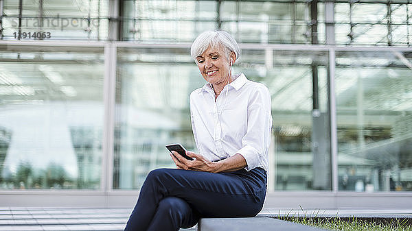 Senior businesswoman sitting outside with smartphone and earbuds