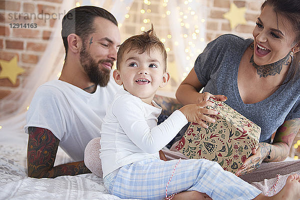 Happy boy opening Christmas present with his parents in bed