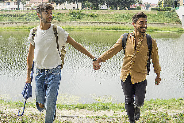 Portrait of young gay couple with backpacks walking hand in hand at riverside
