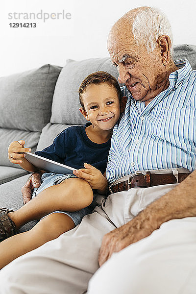 Portrait of happy little boy with digital tablet sitting besides his grandfather on the couch at home