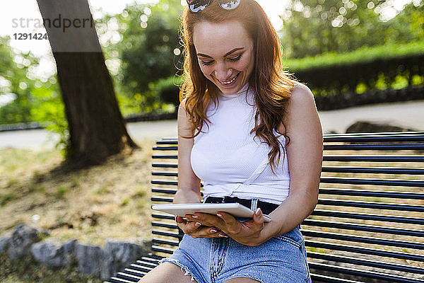 Laughing redheaded woman sitting on bench in park using digital tablet