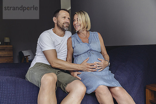 Mature man and his pregnant mature wife sitting on bed touching her belly