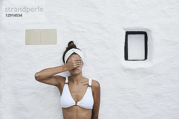 Young woman wearing white bikini top leaning against white wall covering eyes with her hand