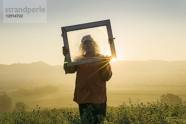 Italy  Tuscany  Borgo San Lorenzo  senior man holding window frame in field at sunrise above rural landscape