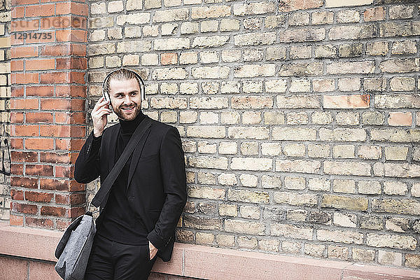 Smiling fashionable young man with headphones leaning against brick wall