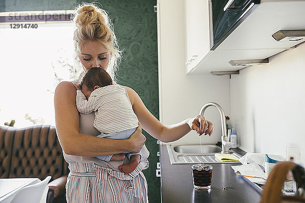 Mother holding newborn baby in kitchen while making tea