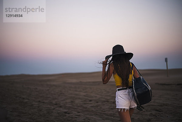 Back view of teenage girl with hat and shoulder bag walking on the beach at sunset