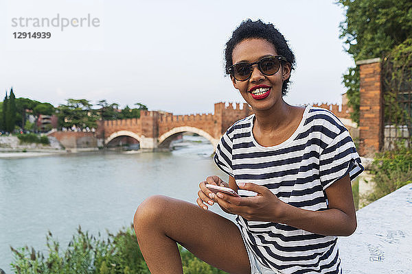 Smiling young woman wearing sunglasses at the riverside using cell phone