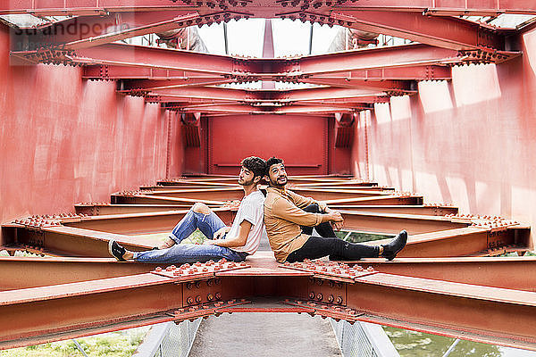 Young gay couple sitting back to back on steel girder of a footbridge