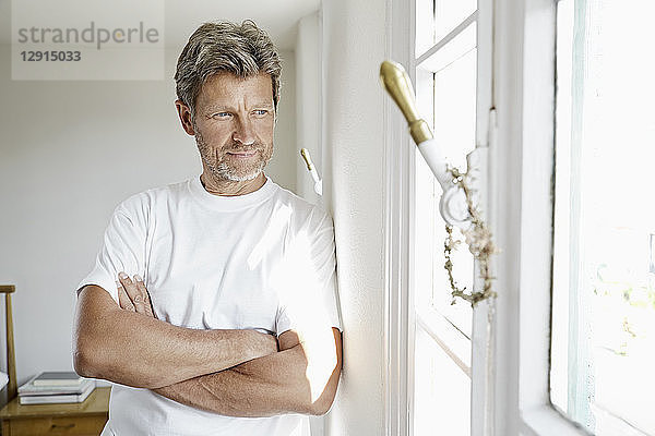 Mature man standing by window with arms crossed