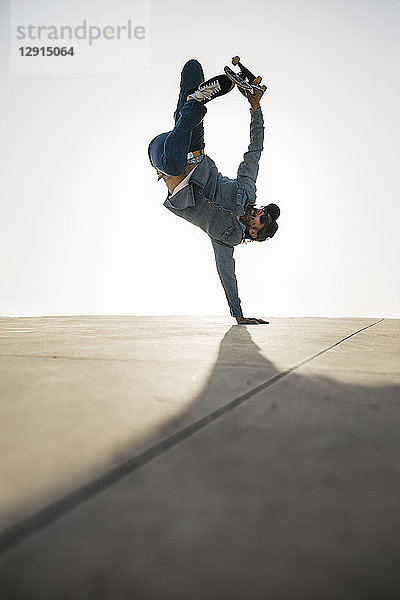 Stylish man in denim outfit showing trick with skate in handstand