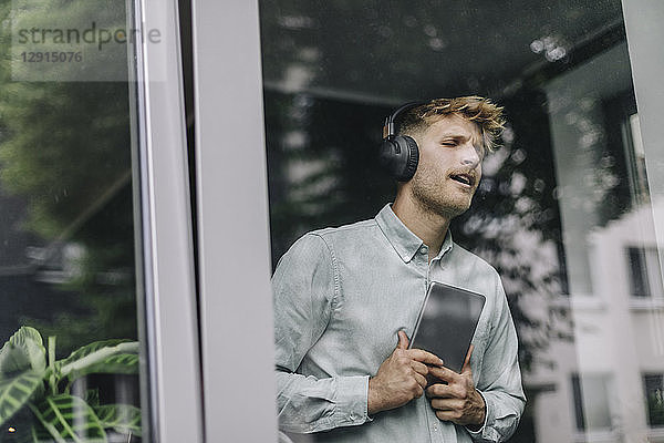 Young man standing at the window  listening music with headphones and digital tablet