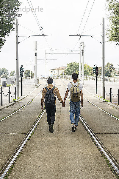 Back view of young gay couple with backpacks walking hand in hand between tracks on the road